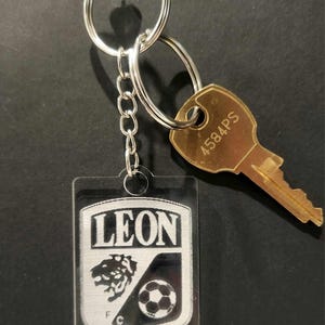 Pode incluir: Um chaveiro de acr&iacute;lico transparente com um logotipo preto e branco de um le&atilde;o e uma bola de futebol. O logotipo diz "LEON" e "F.C.". O chaveiro est&aacute; preso a um chaveiro prateado e uma corrente prateada.