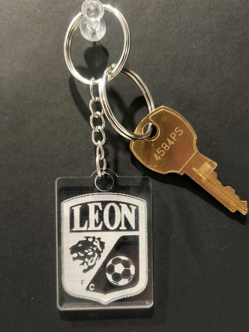 Pode incluir: Um chaveiro de acr&iacute;lico transparente com um logotipo preto e branco de um le&atilde;o e uma bola de futebol. O logotipo diz "LEON" e "F.C.". O chaveiro est&aacute; preso a um chaveiro prateado e uma corrente prateada.