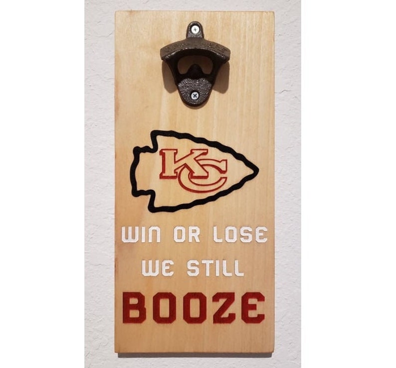 May include: A wooden bottle opener with a black metal bottle opener attached. The wood is stained a light brown color. The bottle opener has a black and red Kansas City Chiefs logo with the words "Win or Lose We Still Booze" printed below.