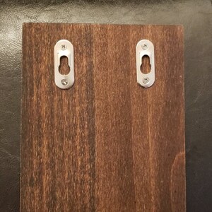 May include: A dark brown wooden sign with two metal hanging hooks on the back. The hooks are silver and have a small hole in the center.