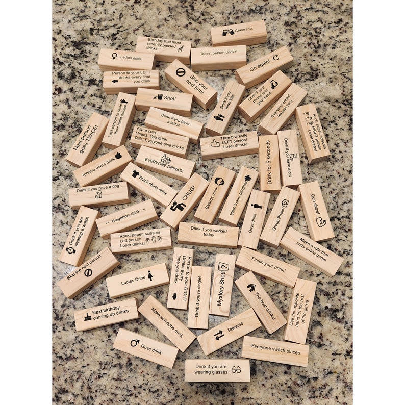 May include: A collection of wooden blocks with various drinking game instructions printed in black. Instructions include "Drink if you have a tattoo," "Everyone drinks," and "Next person goes TWICE!" The blocks are scattered on a speckled surface.