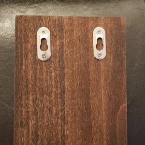 Puede incluir: Una tabla de madera marr&oacute;n oscuro con dos ganchos de colgar de metal plateado. Los ganchos son de forma ovalada con un peque&ntilde;o agujero en el centro.