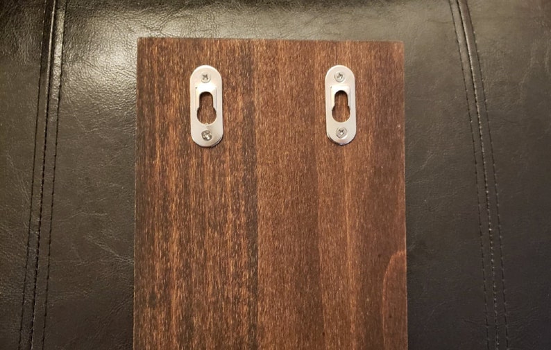 Puede incluir: Una tabla de madera marr&oacute;n oscuro con dos ganchos de colgar de metal plateado. Los ganchos son de forma ovalada con un peque&ntilde;o agujero en el centro.