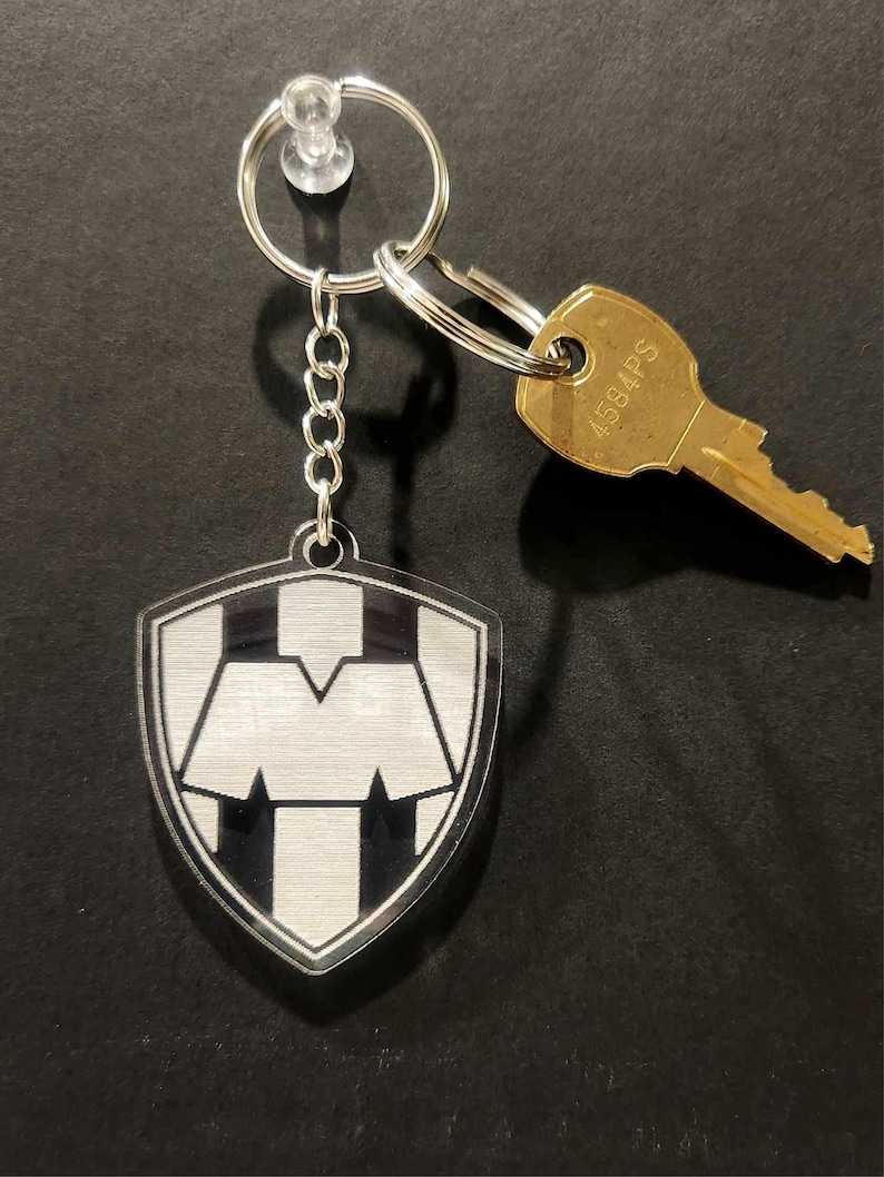 Pode incluir: Um chaveiro em forma de escudo prateado e preto com uma letra M branca no seu interior. O chaveiro est&aacute; ligado a uma corrente prateada e um porta-chaves prateado com uma chave dourada.