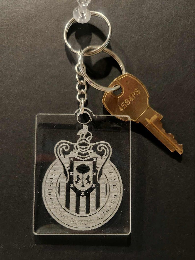 Pode incluir: Um chaveiro de acr&iacute;lico transparente com um logotipo preto e branco de um bras&atilde;o com o texto "Club Deportivo Guadalajara" ao redor. O chaveiro est&aacute; preso a uma corrente prateada e uma chave dourada com o n&uacute;mero "4584PS" nela.