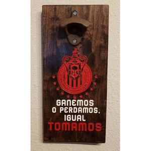 Puede incluir: Un abridor de botellas de madera con un logotipo rojo y blanco de un equipo de f&uacute;tbol. El logotipo dice "Club Deportivo Guadalajara" y tiene una calavera y huesos cruzados en el centro. El texto debajo del logotipo dice "Ganemos o Perdamos, Igual Tomamos".