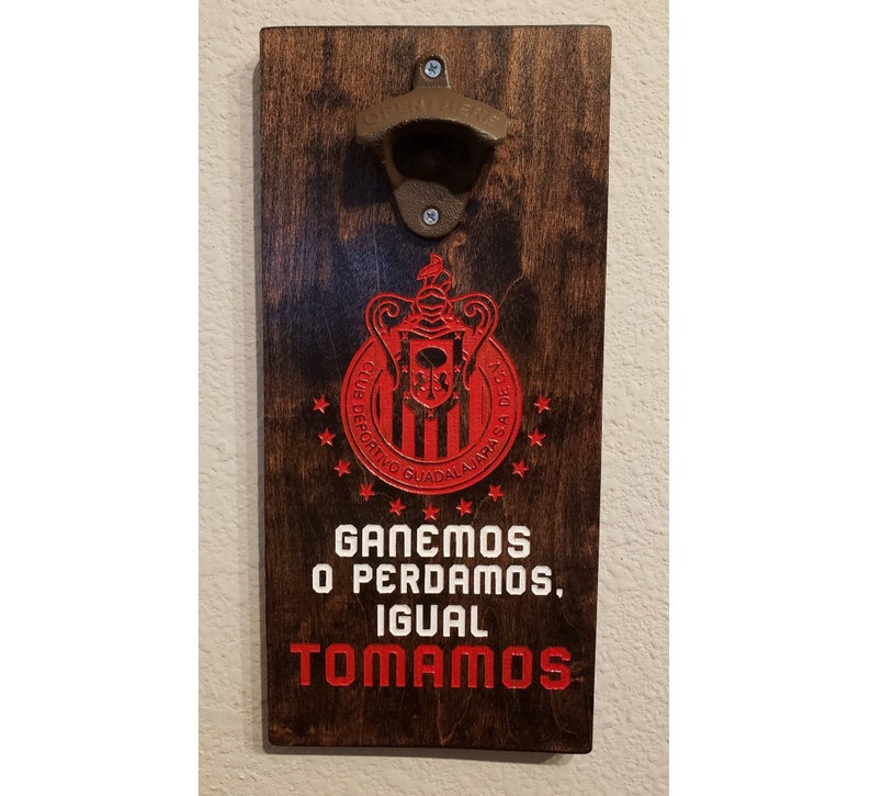 Puede incluir: Un abridor de botellas de madera con un logotipo rojo y blanco de un equipo de f&uacute;tbol. El logotipo dice "Club Deportivo Guadalajara" y tiene una calavera y huesos cruzados en el centro. El texto debajo del logotipo dice "Ganemos o Perdamos, Igual Tomamos".
