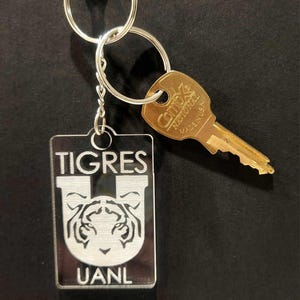 Pode incluir: Um chaveiro prateado com um chaveiro de acr&iacute;lico transparente com um logotipo de tigre branco e o texto "TIGRES UANL".