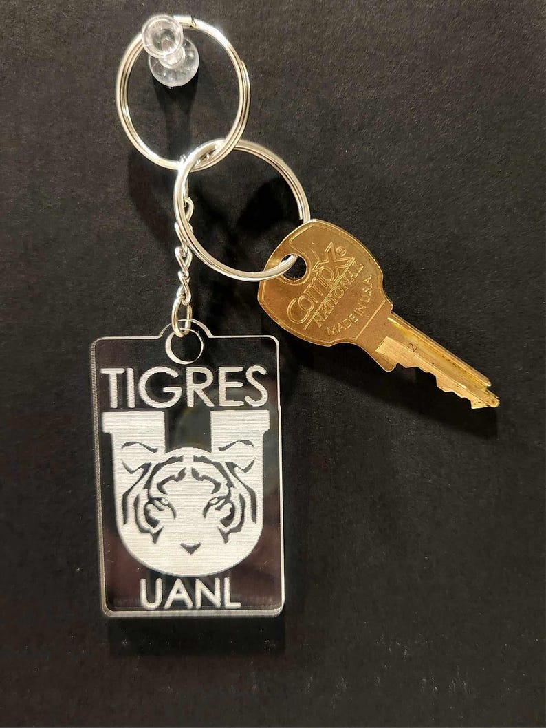 Pode incluir: Um chaveiro prateado com um chaveiro de acr&iacute;lico transparente com um logotipo de tigre branco e o texto "TIGRES UANL".