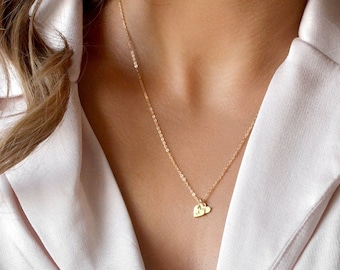 Puede incluir: Un collar de cadena dorada delicada con dos pequeños dijes en forma de corazón. Cada dije está grabado con una letra individual, específicamente los caracteres A y J. El collar se lleva alrededor del cuello sobre una camisa de cuello clara.