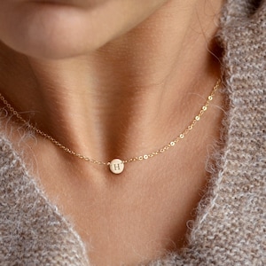 May include: A delicate gold-filled necklace with a small, round pendant bearing the letter "H". The pendant is suspended from a fine chain, offering a minimalist and elegant aesthetic. The necklace is displayed against a neutral backdrop.
