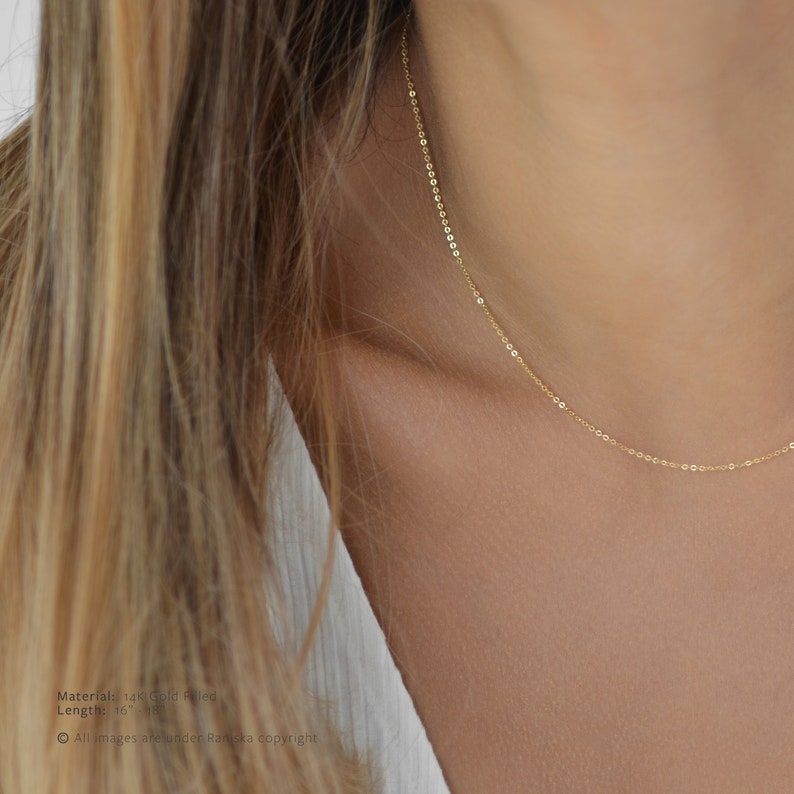 May include: A delicate gold-filled necklace with a fine chain is draped around a person's neck. The necklace is a simple, elegant design, perfect for layering or wearing alone. The chain is approximately 16 inches long.