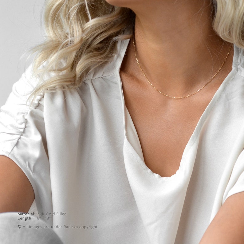 May include: A delicate gold-filled necklace with a subtle, minimalist design. The necklace is shown against a white blouse. The image text indicates the necklace is 16-18 inches long and made of 14K gold filled material.