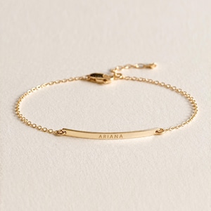 May include: A gold-toned chain bracelet featuring a thin, slightly curved horizontal bar at the center. The bar is engraved with the name ARIANA in capital letters. The bracelet includes a delicate link chain and a small lobster claw clasp.