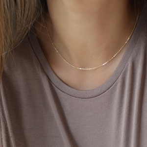 May include: A delicate gold necklace with a subtle design. The necklace features a thin chain with small, evenly spaced circular accents. The jewelry is displayed against a neutral-toned background, highlighting the necklace's simple elegance.