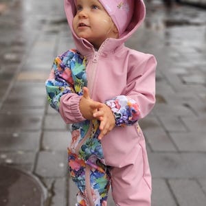 May include: A child's pink hooded jumpsuit with a floral pattern on the sleeves and legs. The jumpsuit has a zipper closure and is paired with a matching pink hat. The child is wearing light green shoes.