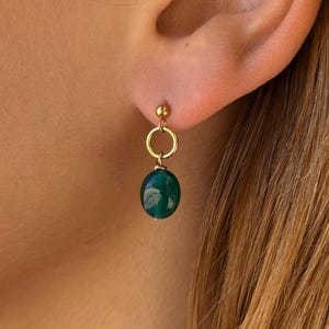Op de afbeelding: Een goudkleurige oorbel met een kleine ronde knop verbonden aan een dunne gouden ring, waaraan een gladde, ovaalvormige donkergroene edelsteen hangt. De oorbel heeft een eenvoudig hangend ontwerp en is afgewerkt met een gepolijste metalen glans.