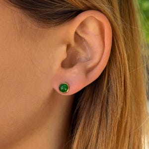 Peut inclure: Une petite boucle d'oreille à clou ronde ornée d'une pierre précieuse verte lisse et vibrante, sertie dans une monture en métal doré poli. La boucle d'oreille est portée au lobe, présentant un design épuré et une couleur verte vive.