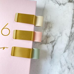May include: A pink notebook with three gold-coloured metal page markers. Each marker holds a different coloured ribbon: cream, pink, and green. The number 6 is printed in gold on the notebook.