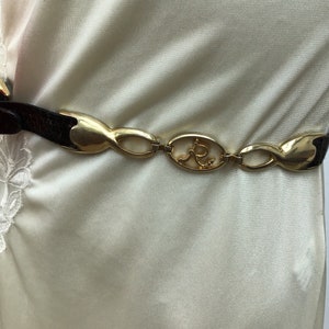 Peut inclure: Une ceinture marron et dorée avec une boucle dorée décorative. La ceinture a une sangle en cuir marron foncé texturé et une série de maillons dorés. La boucle présente un design ovale avec la lettre "R" stylisée.
