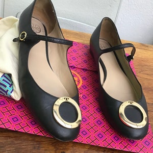 May include: A pair of black leather Mary Jane flats with gold-tone hardware. The shoes feature a round gold buckle and a strap across the top of the foot. The flats are displayed on a pink and purple patterned box.