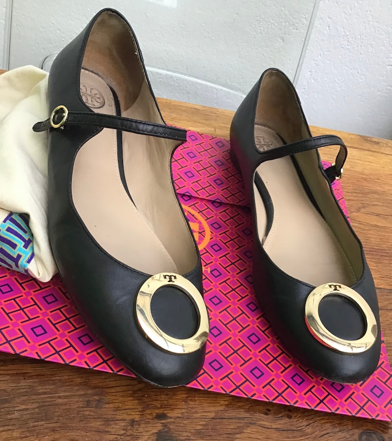 May include: A pair of black leather Mary Jane flats with gold-tone hardware. The shoes feature a round gold buckle and a strap across the top of the foot. The flats are displayed on a pink and purple patterned box.