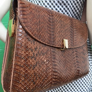 Puede incluir: Un bolso de hombro de piel de serpiente marrón con cierre de broche dorado. El bolso tiene cierre de solapa y una correa larga.