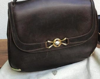 Könnte beinhalten: Eine braune Gucci-Schultertasche aus Leder mit einer goldfarbenen Schnalle und einem Klappverschluss. Die Tasche liegt auf einer weißen Oberfläche mit dem Gucci-Logo.