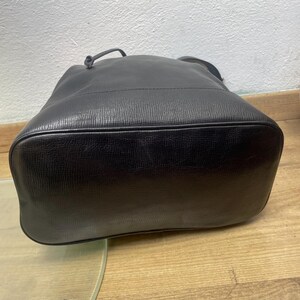 May include: Black leather bucket bag with a textured surface. The bag has a drawstring closure and a single handle. The bottom of the bag is rounded. The bag is sitting on a wooden surface.