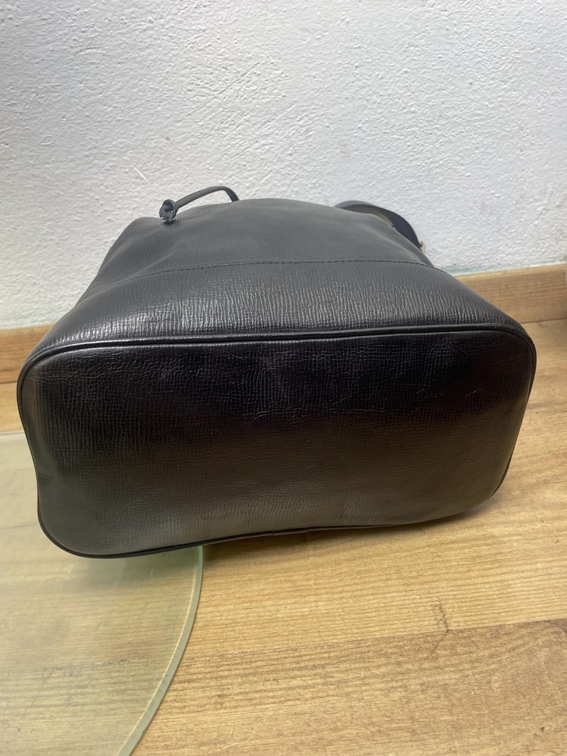May include: Black leather bucket bag with a textured surface. The bag has a drawstring closure and a single handle. The bottom of the bag is rounded. The bag is sitting on a wooden surface.
