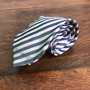 May include: Rolled-up striped tie with navy blue and white stripes, accented with a thin pink stripe. The tie has the word "DSQUARED" embroidered on the end. The tie is made of a smooth fabric.