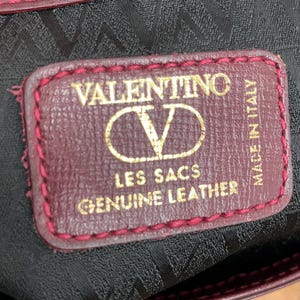 Może przedstawiać: Zbliżenie bordowej sk&oacute;rzanej torby z szytą krawędzią. Etykieta głosi "VALENTINO LES SACS GENUINE LEATHER MADE IN ITALY" złotymi literami. Wnętrze torby jest czarne z powtarzającym się wzorem.