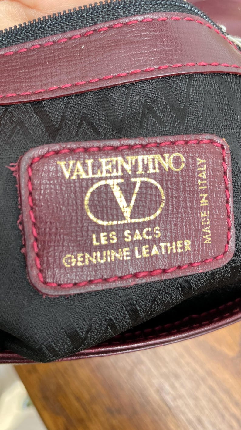 Może przedstawiać: Zbliżenie bordowej sk&oacute;rzanej torby z szytą krawędzią. Etykieta głosi "VALENTINO LES SACS GENUINE LEATHER MADE IN ITALY" złotymi literami. Wnętrze torby jest czarne z powtarzającym się wzorem.