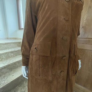 May include: A long, brown suede coat with a button-up front, large collar, and two front pockets. The coat has a classic design and appears to be made of soft suede material. The coat is displayed on a mannequin.
