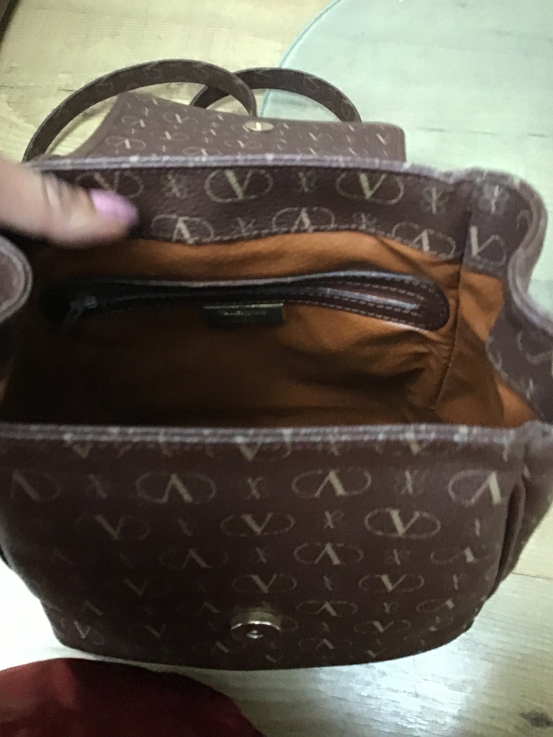 K&ouml;nnte beinhalten: Eine braune Ledertasche mit einem goldenen Verschluss und einem braunen und wei&szlig;en Muster. Die Tasche hat eine Rei&szlig;verschlusstasche im Inneren.