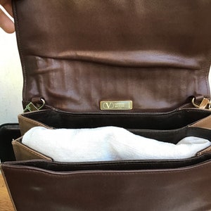 May include: Open view of a brown leather handbag with a gold-colored clasp. The interior is lined with a lighter brown material and contains white padding. A gold-colored metal tag with the brand name is visible.