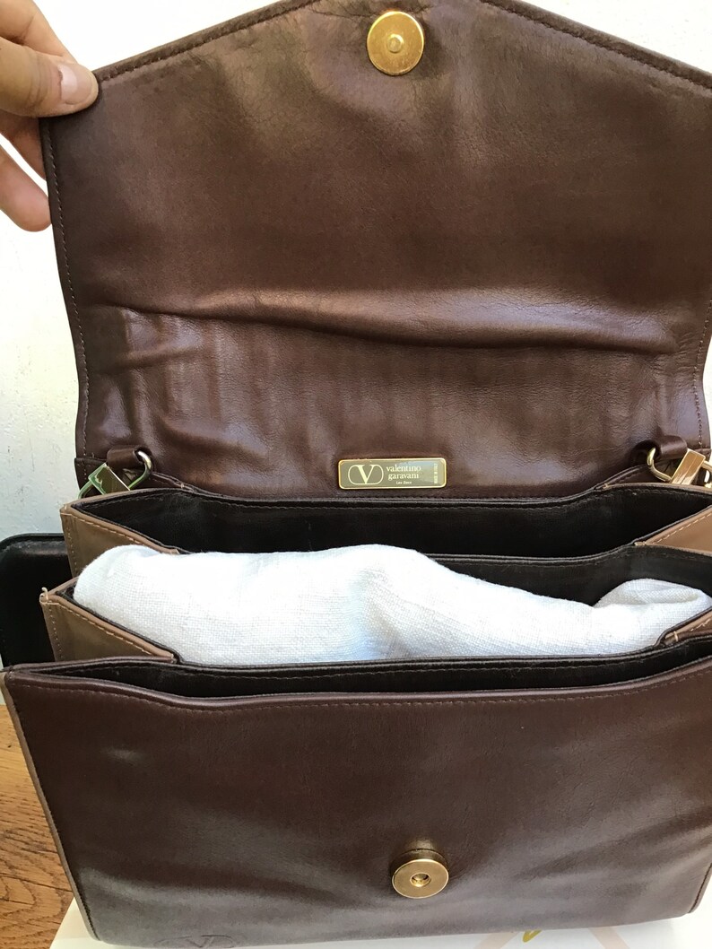 May include: Open view of a brown leather handbag with a gold-colored clasp. The interior is lined with a lighter brown material and contains white padding. A gold-colored metal tag with the brand name is visible.