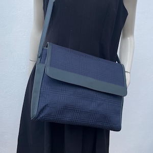 May include: A navy blue crossbody bag with a plaid pattern. The bag has a flap closure and a gray leather strap. The bag is displayed on a mannequin wearing a black dress.