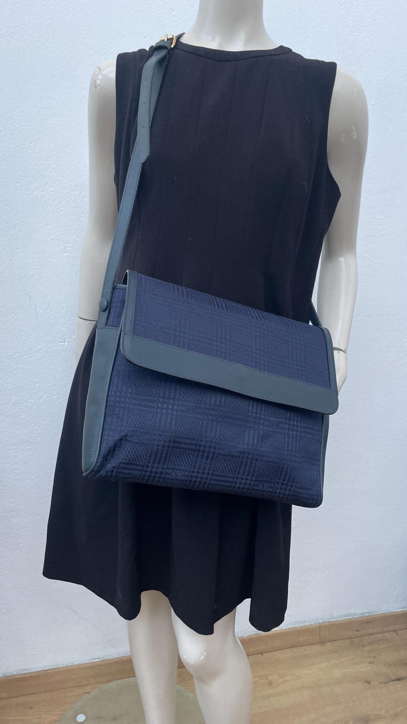 May include: A navy blue crossbody bag with a plaid pattern. The bag has a flap closure and a gray leather strap. The bag is displayed on a mannequin wearing a black dress.