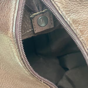 May include: Close-up of a brown leather bag interior. The zipper is partially open, revealing the inside lining. A dark brown snap closure is visible. The leather has a textured appearance.