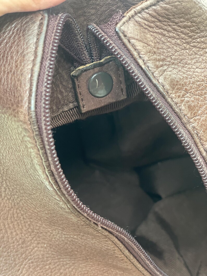 May include: Close-up of a brown leather bag interior. The zipper is partially open, revealing the inside lining. A dark brown snap closure is visible. The leather has a textured appearance.