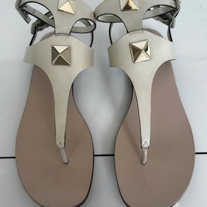May include: A pair of off-white leather sandals with gold-colored square studs. The sandals have a T-strap design and adjustable ankle straps with silver-colored buckles. The footbed is a light tan color.