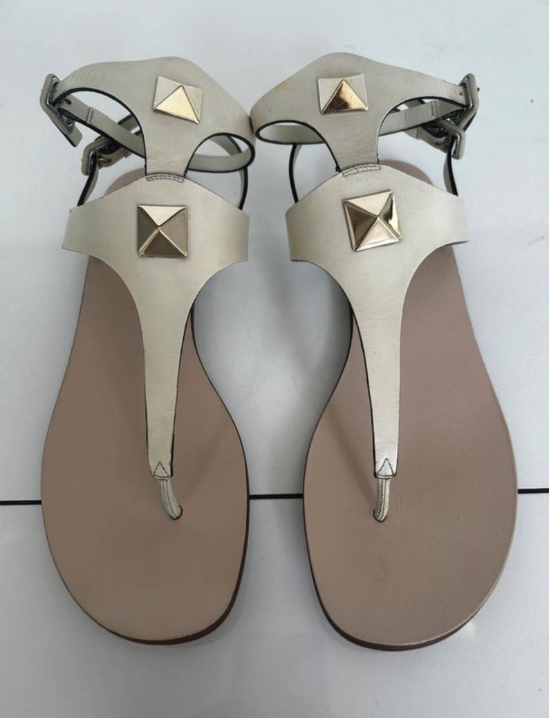 May include: A pair of off-white leather sandals with gold-colored square studs. The sandals have a T-strap design and adjustable ankle straps with silver-colored buckles. The footbed is a light tan color.
