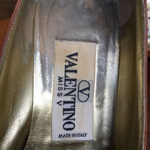 May include: Close-up of the interior of a brown leather shoe. The insole is gold with a white label that reads "VALENTINO MISS V MADE IN ITALY" in black text. The shoe's interior shows signs of wear.