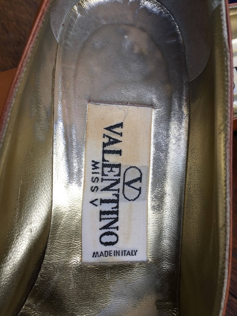 May include: Close-up of the interior of a brown leather shoe. The insole is gold with a white label that reads "VALENTINO MISS V MADE IN ITALY" in black text. The shoe's interior shows signs of wear.