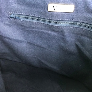 May include: Interior view of a navy blue tote bag. A silver zipper runs horizontally across the top of the bag. A small rectangular tag with the letter "V" is visible. The bag's lining is a solid navy blue fabric.