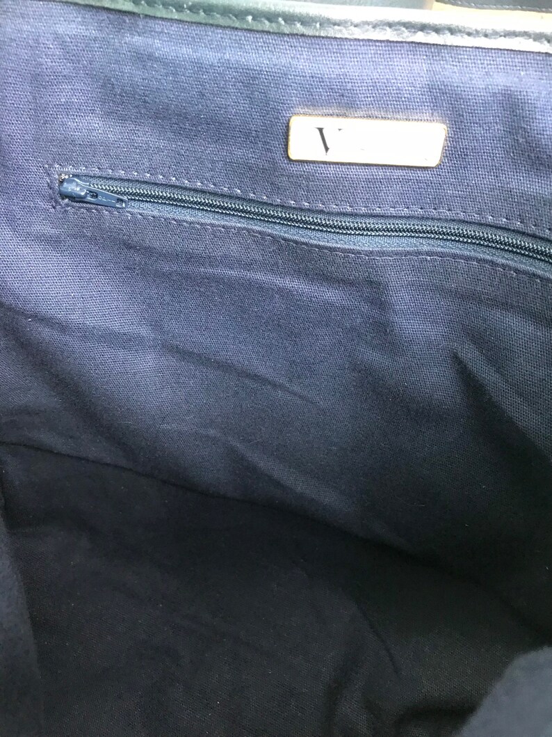 May include: Interior view of a navy blue tote bag. A silver zipper runs horizontally across the top of the bag. A small rectangular tag with the letter "V" is visible. The bag's lining is a solid navy blue fabric.