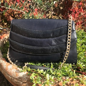 Pode incluir: Uma clutch azul marinho com uma corrente dourada. A bolsa apresenta um padrão texturizado, semelhante à pele de cobra, na frente. A bolsa está em um vaso de terracota com folhagem verde.