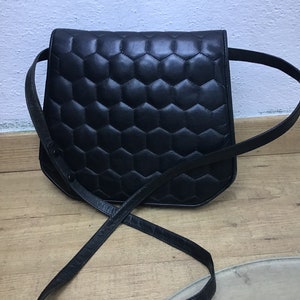 May include: A black leather shoulder bag with a geometric, honeycomb-like quilted pattern. The bag has a long, thin strap and a curved shape. The bag is on a wooden surface.