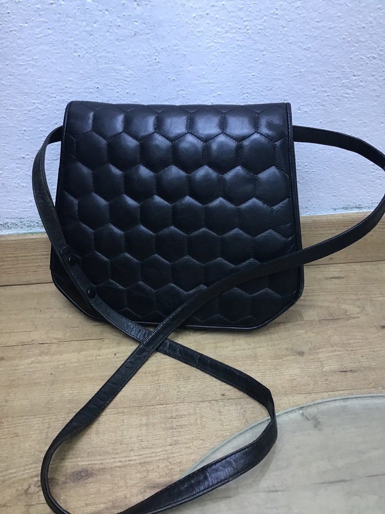 May include: A black leather shoulder bag with a geometric, honeycomb-like quilted pattern. The bag has a long, thin strap and a curved shape. The bag is on a wooden surface.